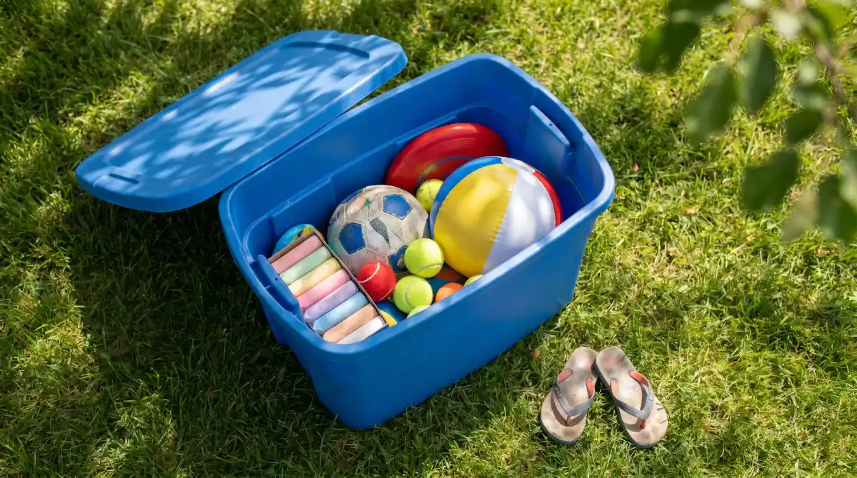 Open outdoor toy bin filled with balls and outdoor games on a sunny lawn